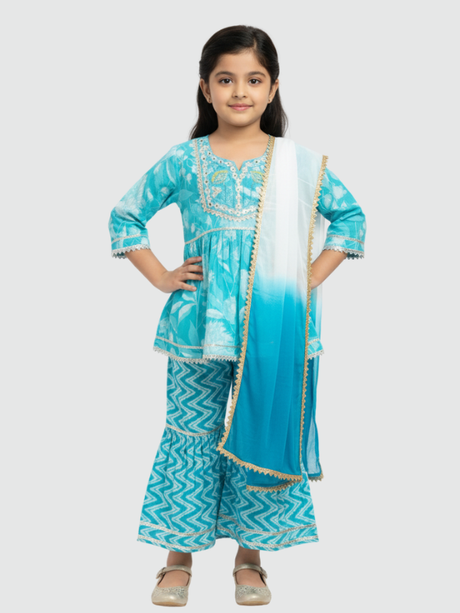 Full-length image of a young girl wearing a turquoise and blue tie-dye ethnic set with an embroidered sleeveless peplum top, tiered gharara pants with a zig-zag print, and a white-to-blue ombre dupatta.