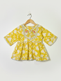 Front view of a girls' yellow and white floral-printed peplum top (kurti) with 3/4 sleeves and a heavily embroidered neckline detailed with mirror work, hanging on a white background.