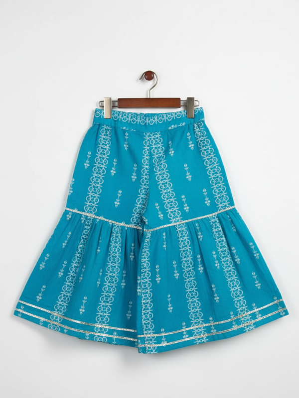 Full-length image of a young girl wearing a turquoise and blue floral-printed ethnic set with a flared kurta, tiered gharara pants, and a vibrant pink and blue ombre chiffon dupatta.