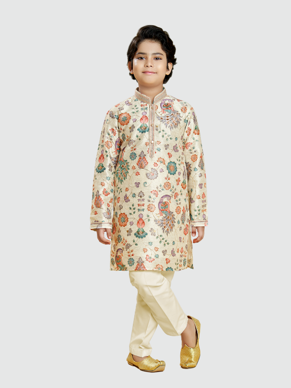 Young boy posing in a full-sleeve cream/light gold kurta with an intricate, multi-color woven peacock and floral pattern, paired with cream pajamas and bright golden traditional footwear.